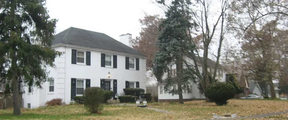 A white-painted two-storey colonial house with black shutters, clipped hedges and mature conifers, under overcast light