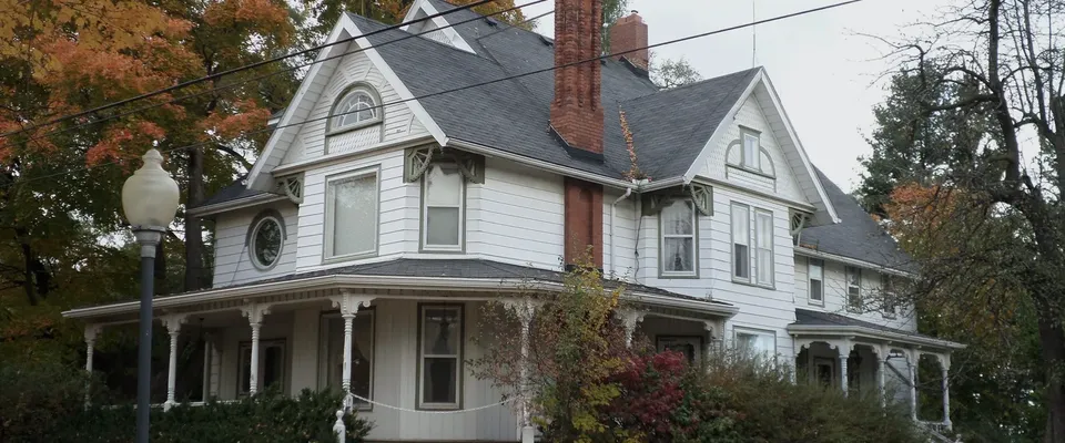 A large white Queen Anne house with a wraparound porch and turned columns, under autumn maples