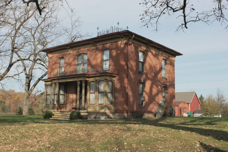 A nineteenth-century brick farmhouse with a covered porch, photographed from the roadside in soft daylight