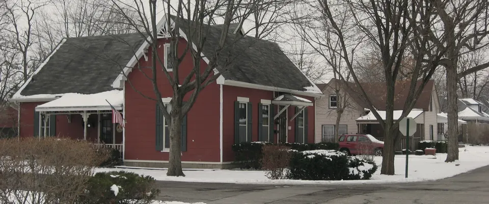 A frame house with a covered porch set back behind a lawn on a quiet residential road