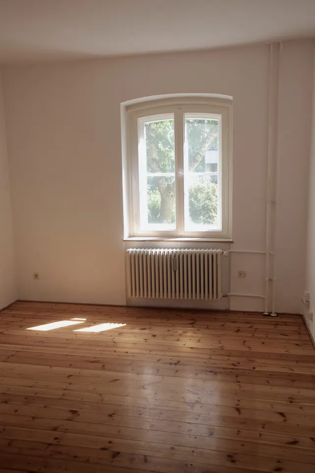 An empty upstairs room with bare floorboards, a tall window and a cast-iron radiator.